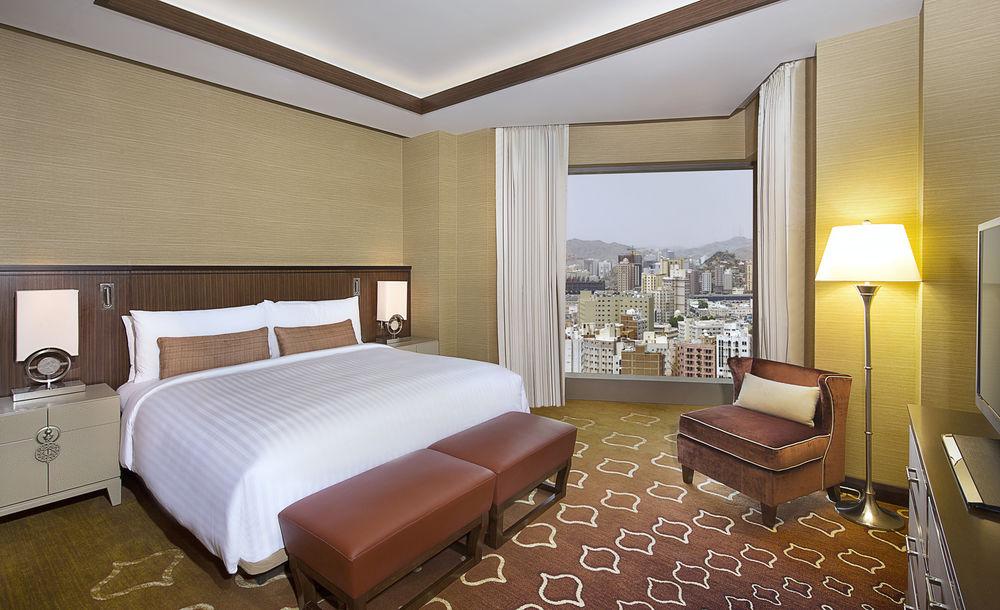 Jabal Omar Marriott Hotel Makkah La Meca Exterior foto The photo shows a modern hotel room featuring a large bed with white linens and decorative cushions. There are two brown leather footstools at the foot of the bed. Next to the bed is a stylish armchair in a complementary color. The room has large win