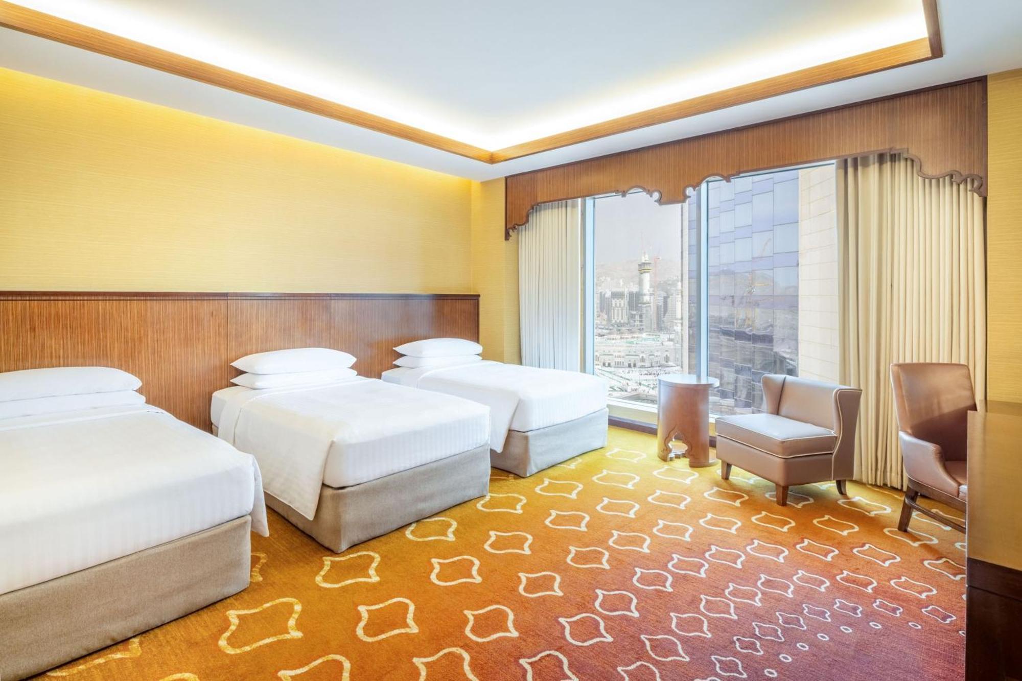 Jabal Omar Marriott Hotel Makkah La Meca Exterior foto The photo depicts a hotel room featuring three beds arranged in a neat manner. The walls are painted a soft yellow, and there is a decorative wooden panel behind the beds. The carpet has a unique orange design, contributing to a warm atmosphere. Larg